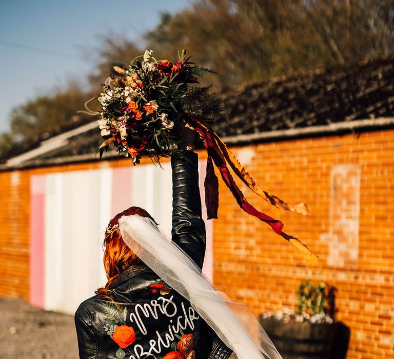 Bride wearing a leather jacket cover up with personalised details and roses holding up her red and orange bouquet 