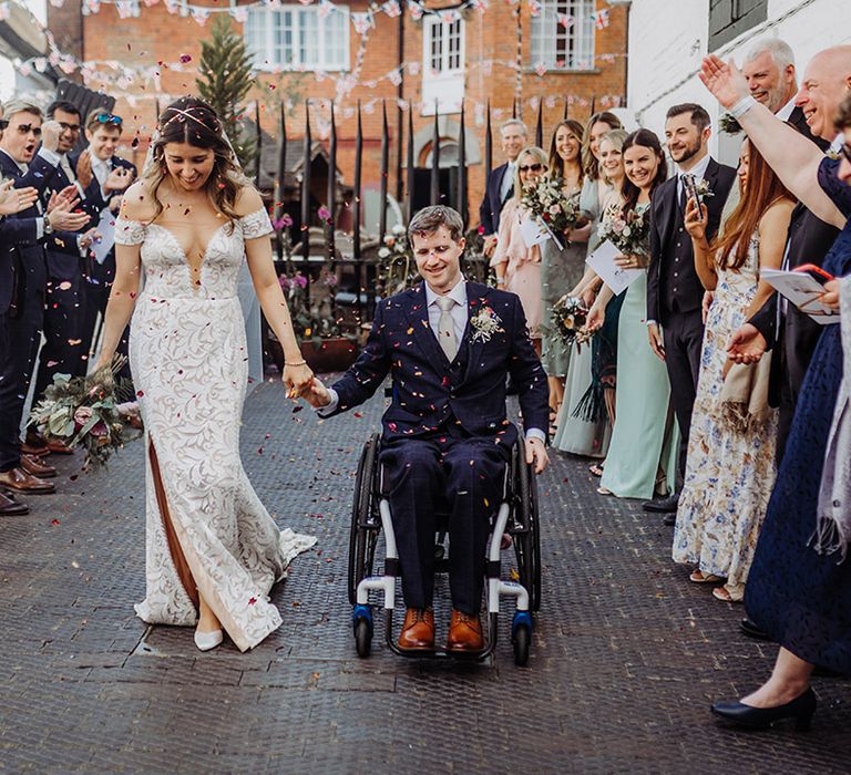 Bride in boho lace off the shoulder wedding dress with gold hair piece holding hands with the groom in a dark blue suit and gold tie as they have their confetti moment