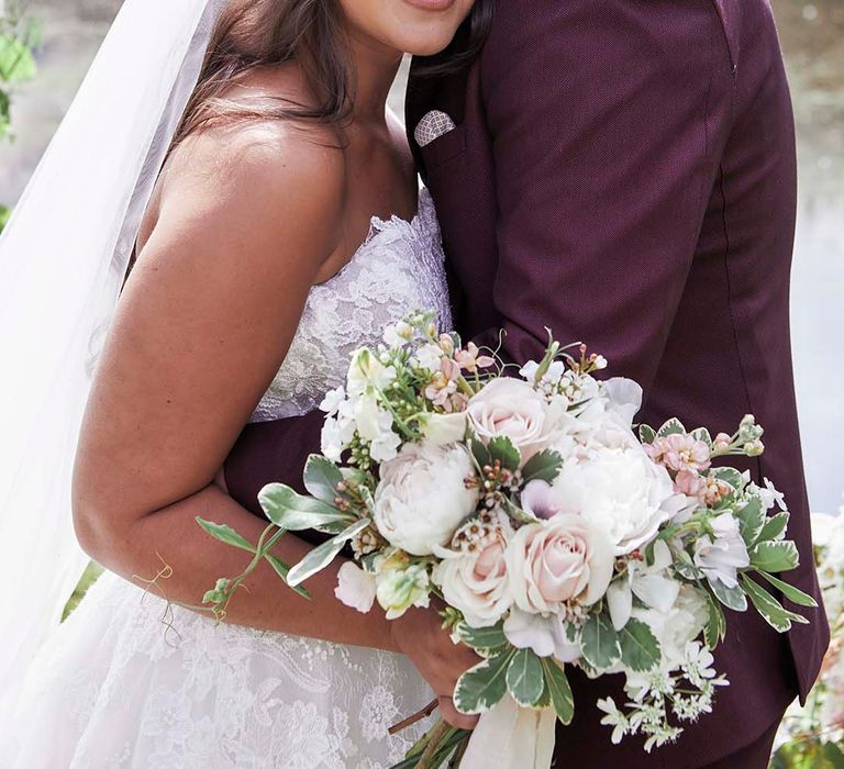 Groom in a burgundy wedding suit embracing his bride in a strapless lace wedding dress with long wavy hair and veil 