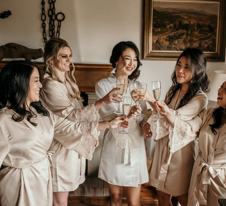 Bride wears short white silk dressing gown complete with lace sleeves and holds champagne glass as bridesmaids wearing matching dressing gowns in champagne colour