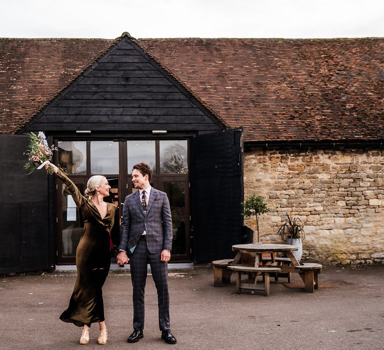 Bride wears ankle length velvet wedding dress whilst holding pastel floral bouquet and holding the hang of her groom in tartan suit 