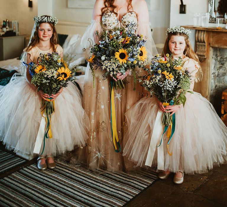 Bride wears pink starry wedding dress with sheer off-the-shoulder sleeves whilst stood with flower girls in pink ballerina dresses all holding sunflower bouquets