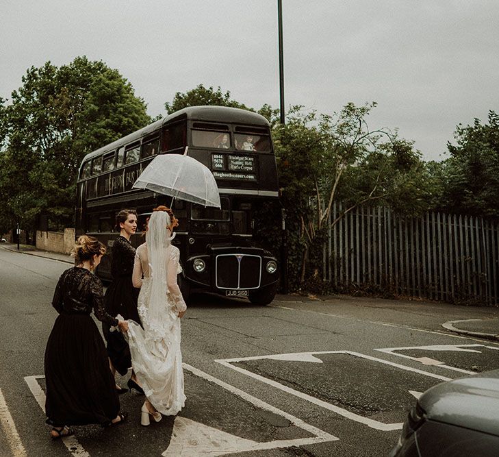 Bride walks with her bridesmaids who wear black bridesmaid dresses and hold see through umbrella above brides head