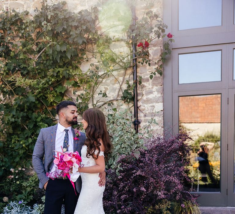 Bride & groom stand outdoors on their wedding day at the Priston Mill as bride holds bright pink floral bouquet 