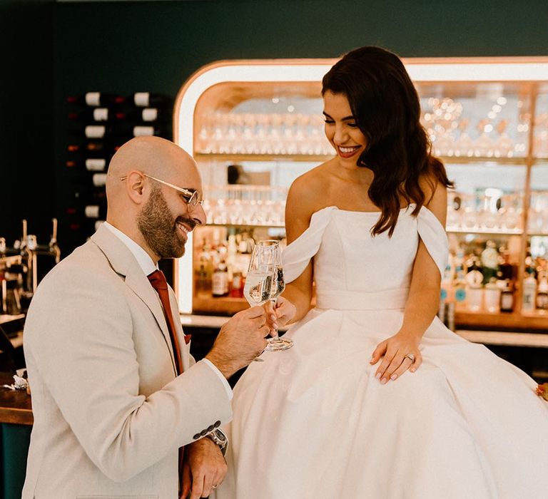 Bride in her off the shoulder wedding dress sits on the bar making a toast with the groom 