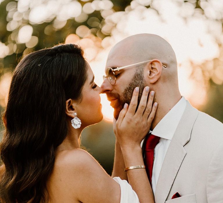 Bride with pearl earrings leaning in to kisses the groom in a pale grey suit and red tie