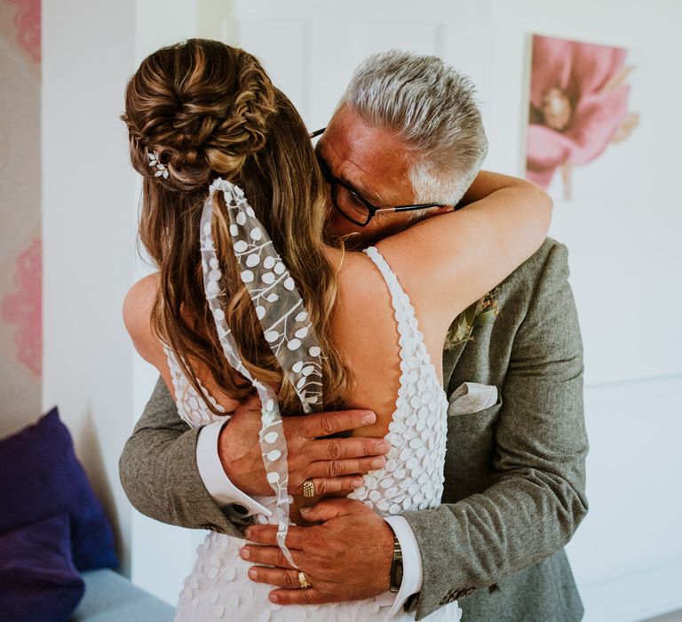 Bride in sleeveless Made With Love Bridal dress with white embellishments and deep plunge back with matching hair ribbon embracing man in grey suit with gold rings