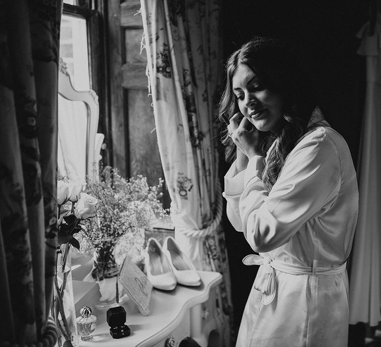 Bride puts in her earrings wearing a white satin robe for her classic country house wedding 