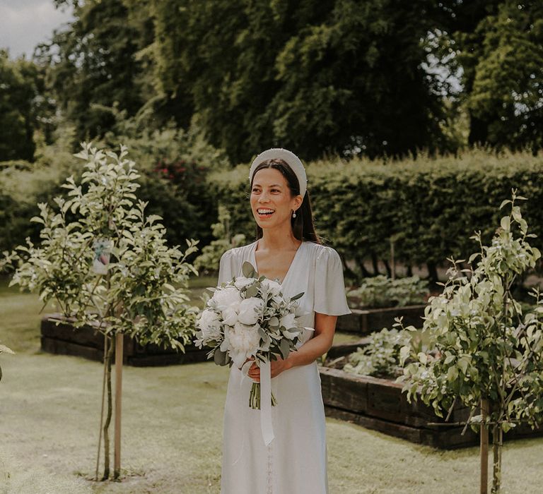 Bride in a button satin wedding dress wearing a chunky pearl wedding headband with a white flower bouquet 