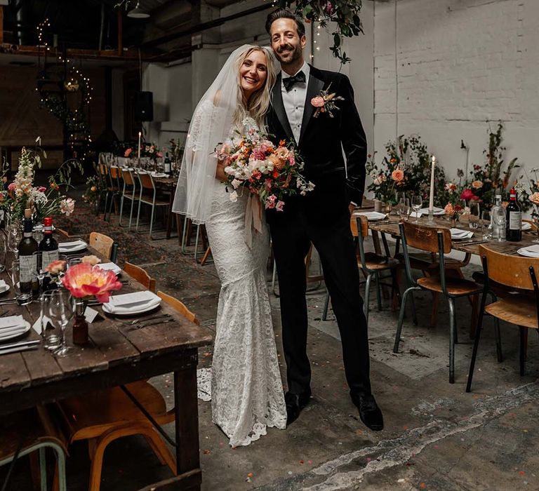 Bride in boho two piece lace bridal outfit with large colourful bouquet and groom in black tux with velvet look bow tie standing in reception room at Hackney Studios