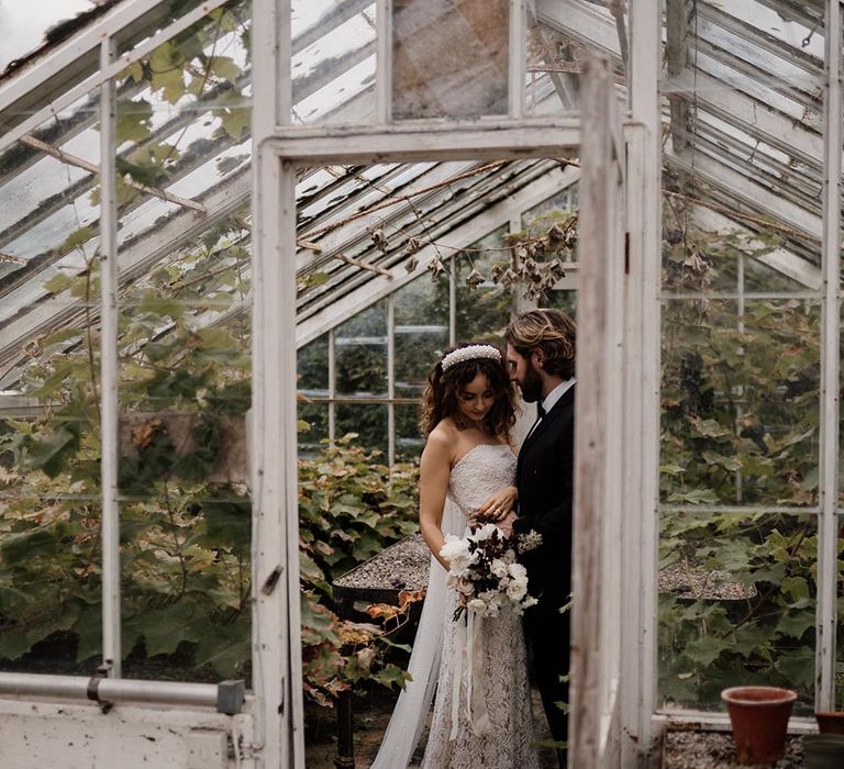 Bride in sleeveless lace wedding dress, pearl headband and black and white bouquet standing with groom in black tux in greenhouse