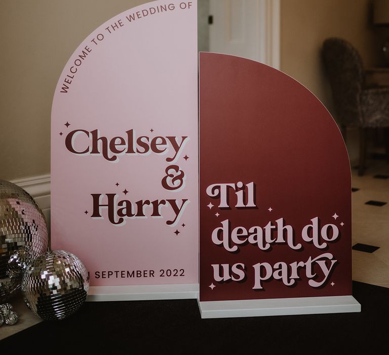 Two toned semi-circle welcome wedding signs in baby pink and brick red with 70's-inspired fonts and graphics and disco ball decor