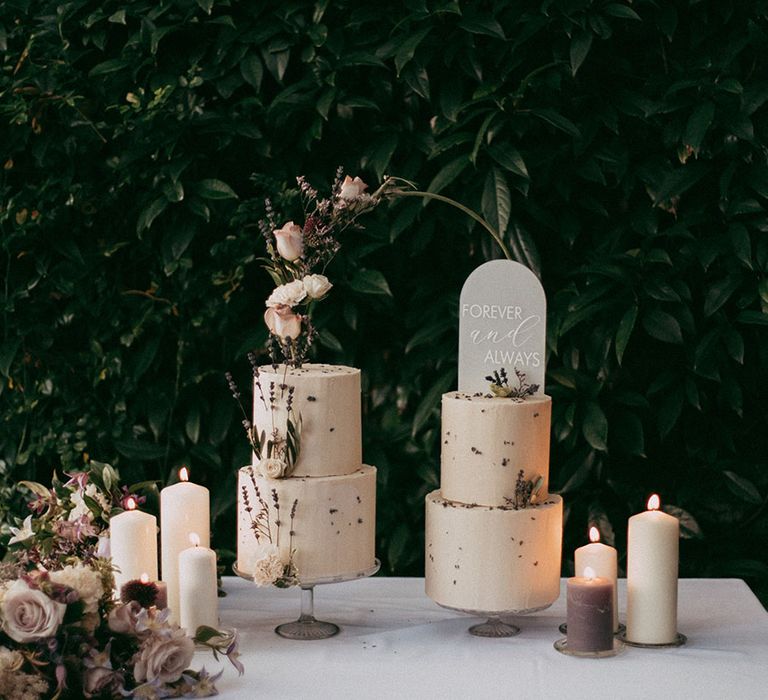 Two tiered buttercream wedding cakes with dried lavender and white rose decorations, white and lavender pillar candles, and neutral toned rose and foliage flower arrangements 