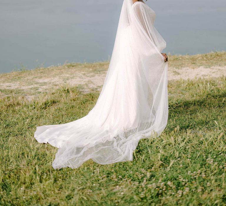 Bride holds pearl detailed veil in front of ocean at destination wedding in Portonovo, Italy