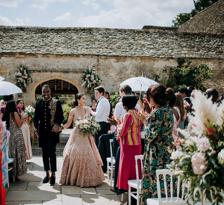 Bride and Groom walk up the aisle holding hands in golden bridal Lehnga and midnight blue and gold Indian groom outfit