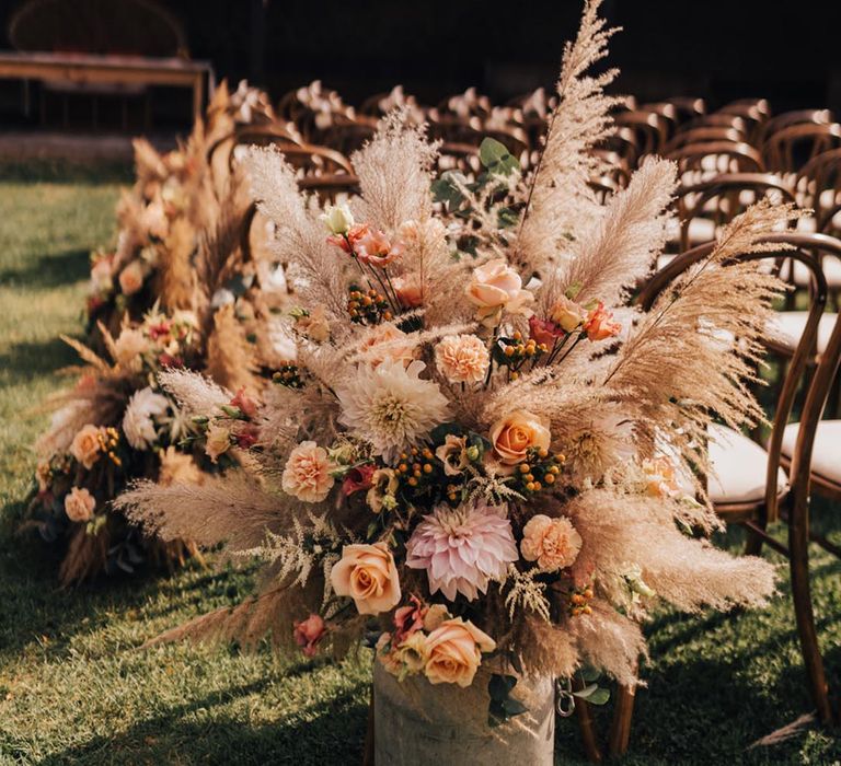 Orange roses, carnations, dahlias and pampas grass in milk churn for outdoor aisle decoration 