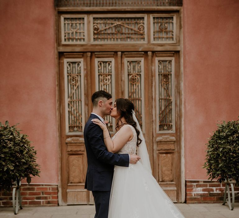 Bride in lace wedding dress and cathedral veil kisses her groom outside Le Petit Chateau 