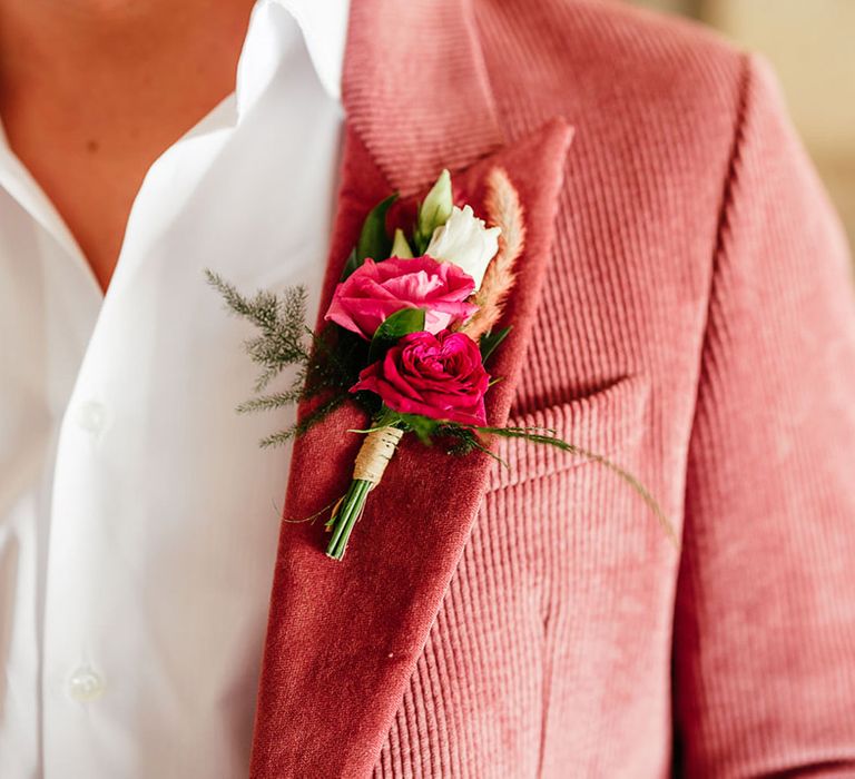 Groom showing off pink and red rose, foliage and dried flower boutonniere on his pink corduroy suit