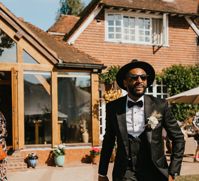 Groom wears three piece suit complete with sunglasses, fedora and white floral buttonhole 