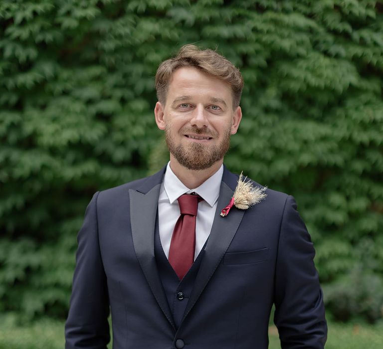 Groom wears three piece suit complete with burgundy tie and pampas grass buttonhole 