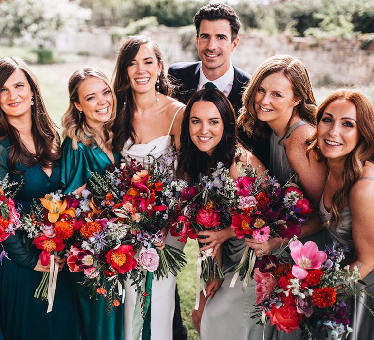 Bridesmaids in mismatched green bridesmaid dresses stand smiling with the bride and groom all holding bright pink, red, and orange wedding bouquets 