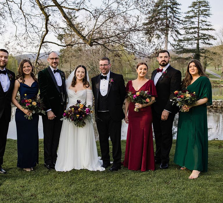 Wedding party photo with the bridesmaid in mismatched jewel tone bridesmaid dresses and the groom and groomsmen in traditional black tie and the bride 