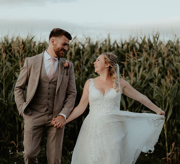 Bride holds her tulle skirt and stands beside her groom in three piece neutral toned suit with pink tie 