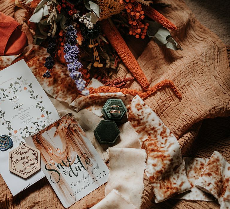 Boho wedding stationery on orange fabric with forest green velvet ring holder, golden wedding rings and colourful dried flowers