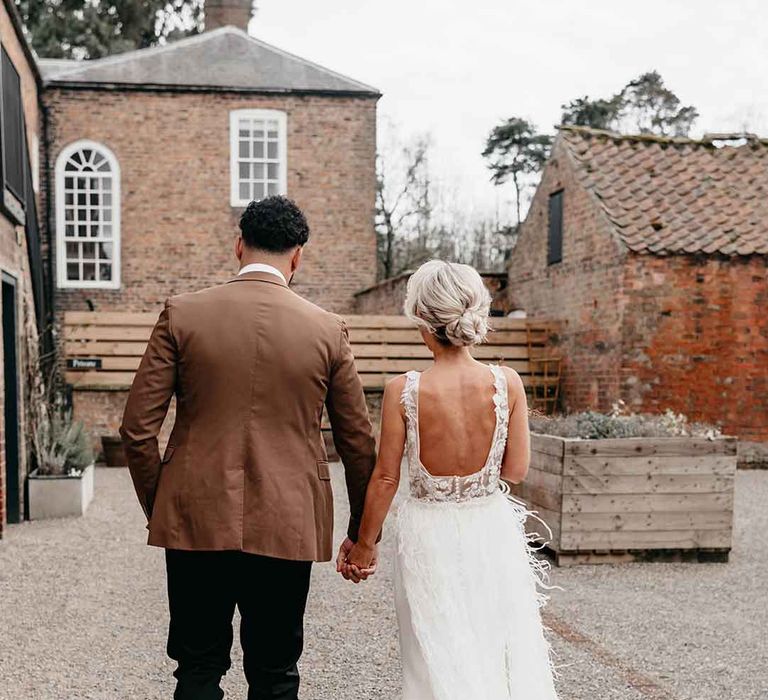 Groom in mocha brown grooms suit and black trousers holding hands with bride wearing Nori Ivy wedding dress with low back and fringe overlay