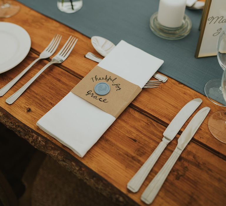 Wooden table with silver cutlery and blue table runner decorated with bud vases of daisies and a white napkin with cardboard holder and wax seal