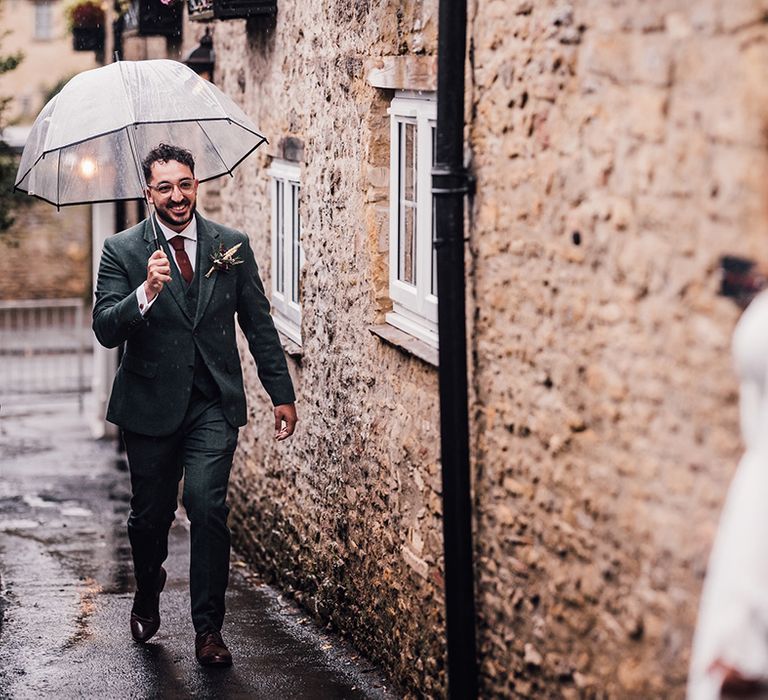 The groom in a dark wedding suit holding an umbrella walks towards the bride in an off the shoulder wedding dress 