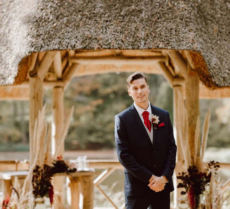 Groom in deep blue grooms blazer, grey waistcoat, red tie, red pocket square and mixed dried flower boutonniere waiting at the alter 