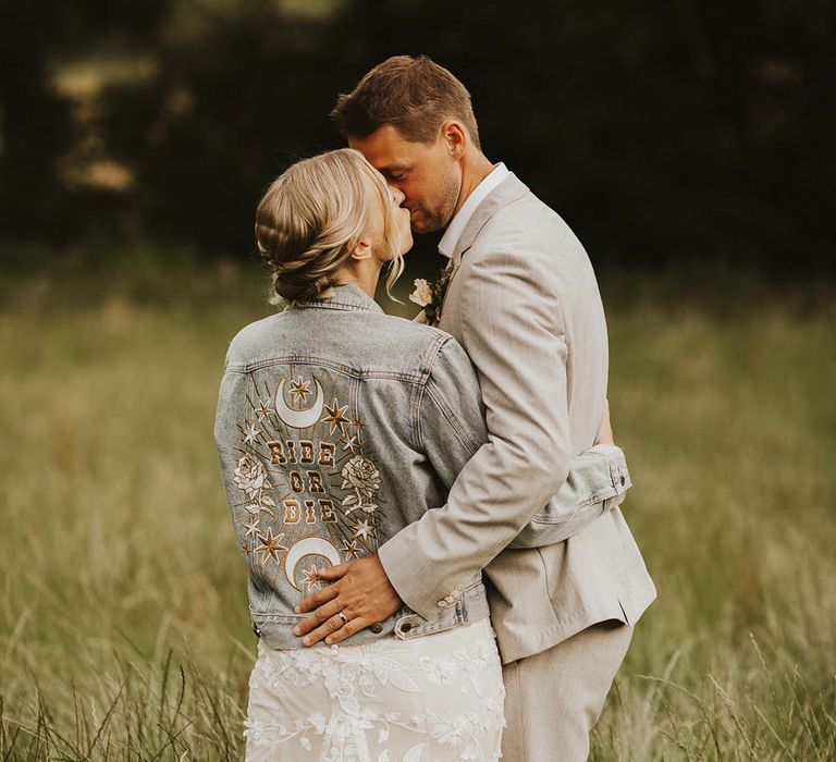 Bride in lace wedding dress and light blue wash denim jacket with 'ride or die' motif kissing the groom in light cream suit for Nancarrow Farm wedding