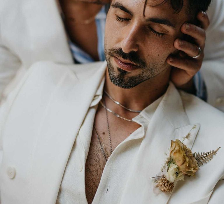 Groom in white blazer, dusky orange garden rose and dried flower boutonniere, white shirt and silver jewellery with groom in white suit and white and blue shirt 