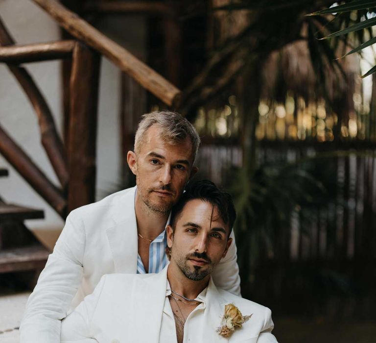 Groom in mens white wedding suit with dusky orange garden rose and dried flower boutonniere, white shirt, white suit trousers and silver jewellery posing with groom in white blazer, light blue and white striped shirt, dusky orange garden rose and dried flower boutonniere, white suit trousers and gold jewellery 