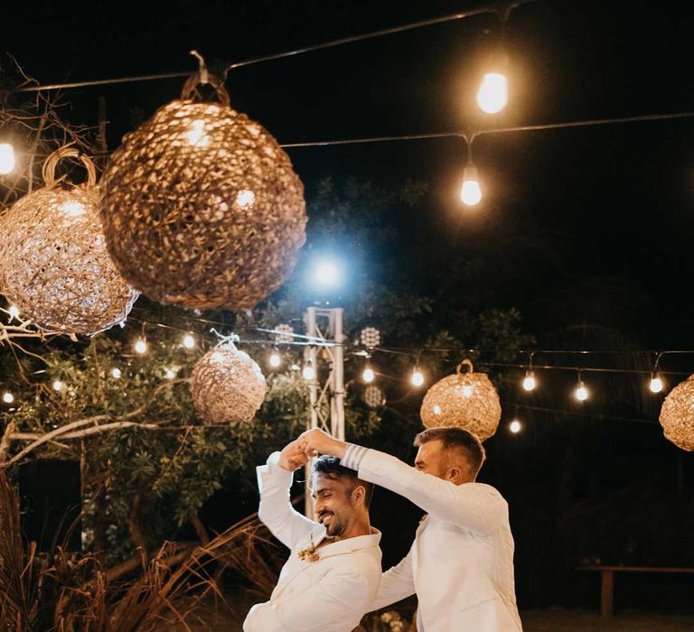 Grooms in matching white grooms suits dancing on DIY hand painted wedding dance floor under twine lampshades and fairy lights 