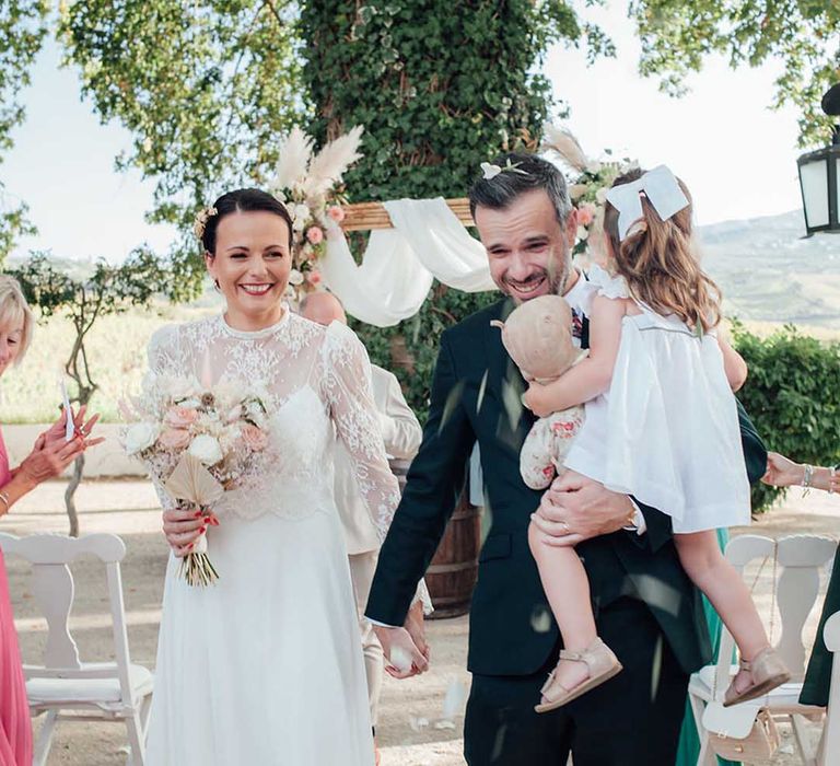 Boho bride in a lace wedding dress and groom in black suit holding his daughter with a hair bow as they walk down the aisle 