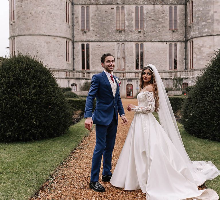 Bride in Eva Lendel wedding dress with long train posing with groom in blue suit at castle wedding 