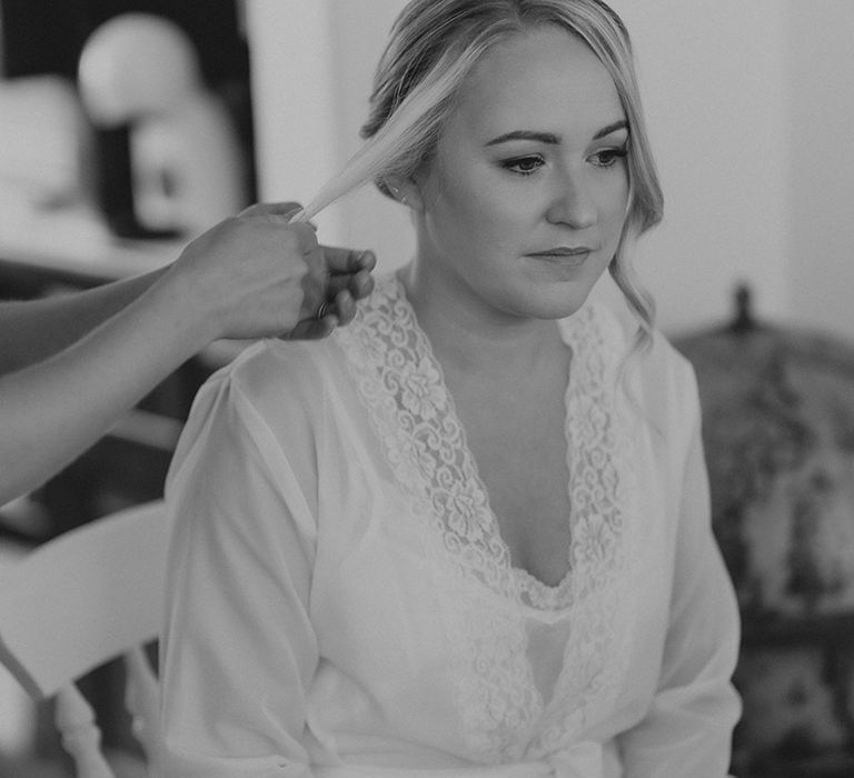 Bride in lace white trim robe sits getting her hair done for the wedding day