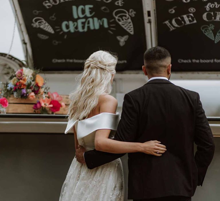 Bride in off the shoulder lace wedding dress standing with groom in classic three piece grooms tuxedo by wedding food truck at Frensham Hall