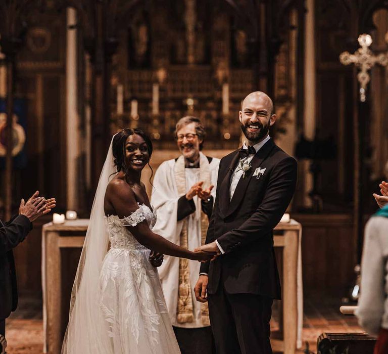 Groom in classic black three piece grooms suit with bow tie and white rose and foliage boutonniere in church holding hands with bride in sweetheart neckline lace wedding dress with detachable off the shoulder sleeves in church wedding 