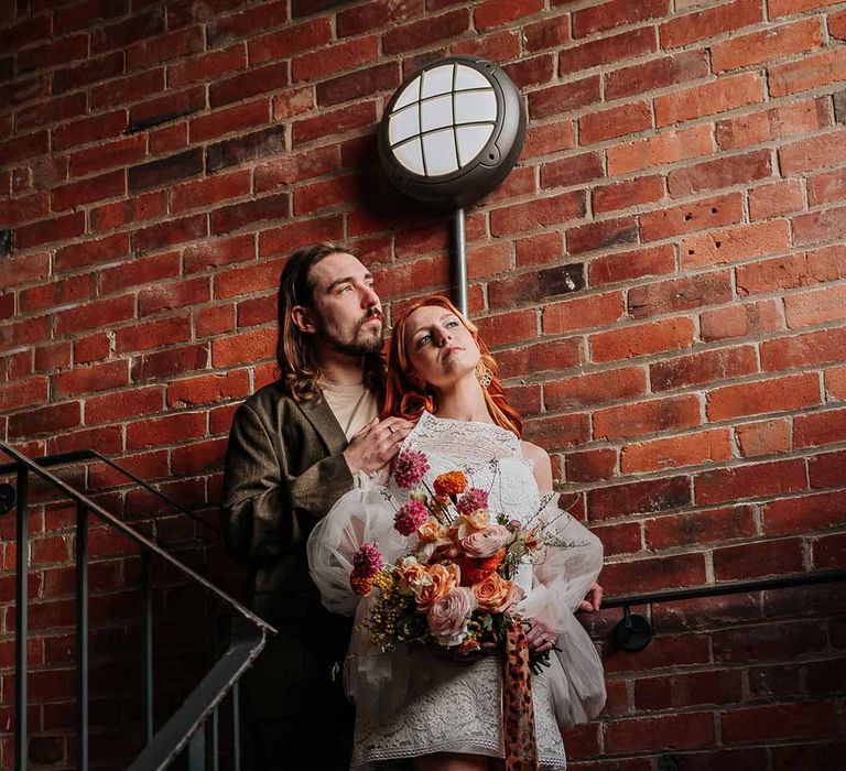 Bride in 70s-inspired boatneck short lace wedding dress with detachable tulle puff sleeves and large platform gold wedding heels holding large colourful mixed flower bridal bouquet with groom in brown blazer, white shirt, grey suit trousers and vans shoes 