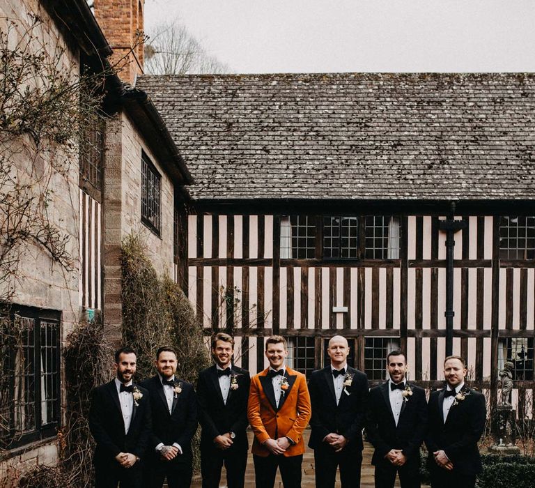 Groomsmen wearing classic black grooms suits with bowties standing with groom in crushed burnt orange velvet grooms blazer, black bowtie and black suit trousers outside Brinsop Court wedding venue