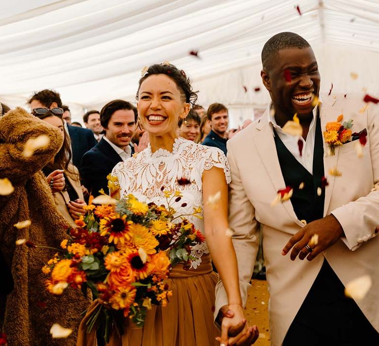 Groom in cream morning suit with forest green grooms waistcoat and trousers, forest green pocket square and orange boutonniere doing colourful dried petal confetti wedding exit at Broadfield Court hand in hand with bride in white mesh lace bridal crop top and yellow wedding skirt