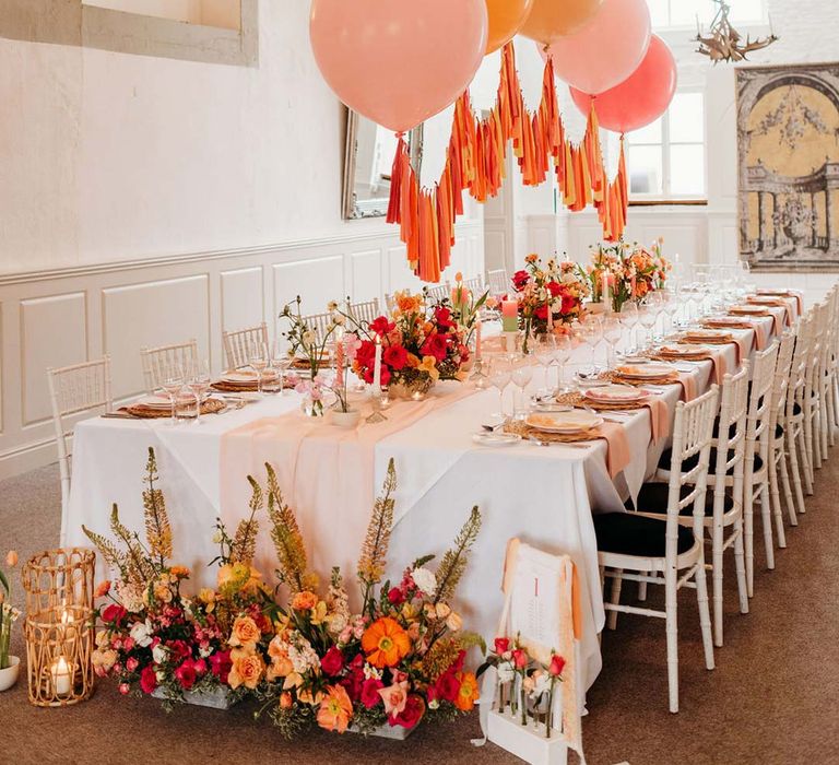 Wedding tablescape with white wedding tablecloth, burlap place mats, tapered candles, pink and orange wedding stationery, and floral wedding table centrepieces with pink and orange garden roses, orange floribunda, yellow ranunculus flowers, foliage and Iceland poppies with red, pink orange and yellow balloons, wedding streamers and fairy lights at Merriscourt wedding venue