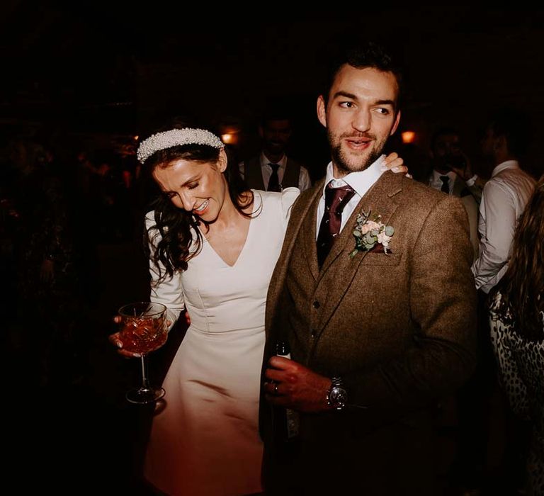 Bride in long sleeve short wedding dress and pearl bridal headband posing with groom in three piece chocolate brown grooms suit 