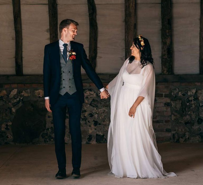 Groom in classic black grooms suit with black tie, grey waistcoat and colourful rose boutonniere standing hand in hand with bride in long sleeve mesh overlay wedding dress and colourful bridal flower crown at Templars Barn