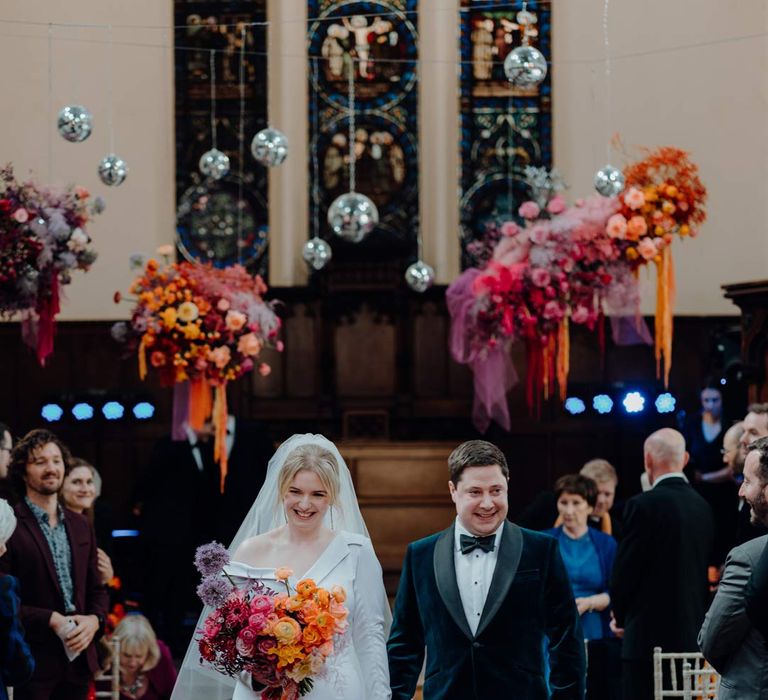 Groom in classic black tuxedo and bowtie walking down the aisle with bride in asymmetrical off shoulder long sleeve satin wedding dress with cathedral length personalised veil holding colourful bridal bouquet with pink garden roses, purple giant alliums, dark red poppies and foliage and their puppy at Fabrica Brighton wedding