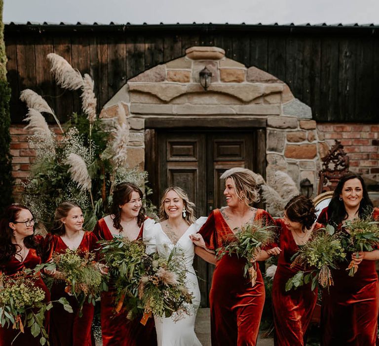 Bridesmaids in matching short sleeve orange velvet bridesmaid dresses walking with the bride holding foliage bouquet 