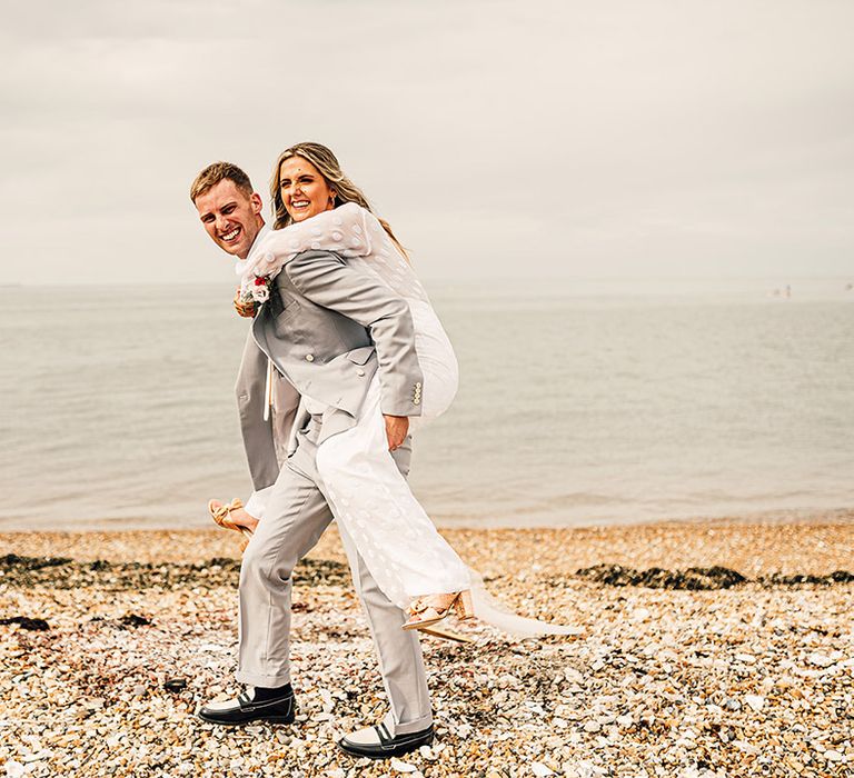 groom in a light grey suit carrying his bride in a jumpsuit on his back as they walk along the beach at their East Quay Whitstable wedding 